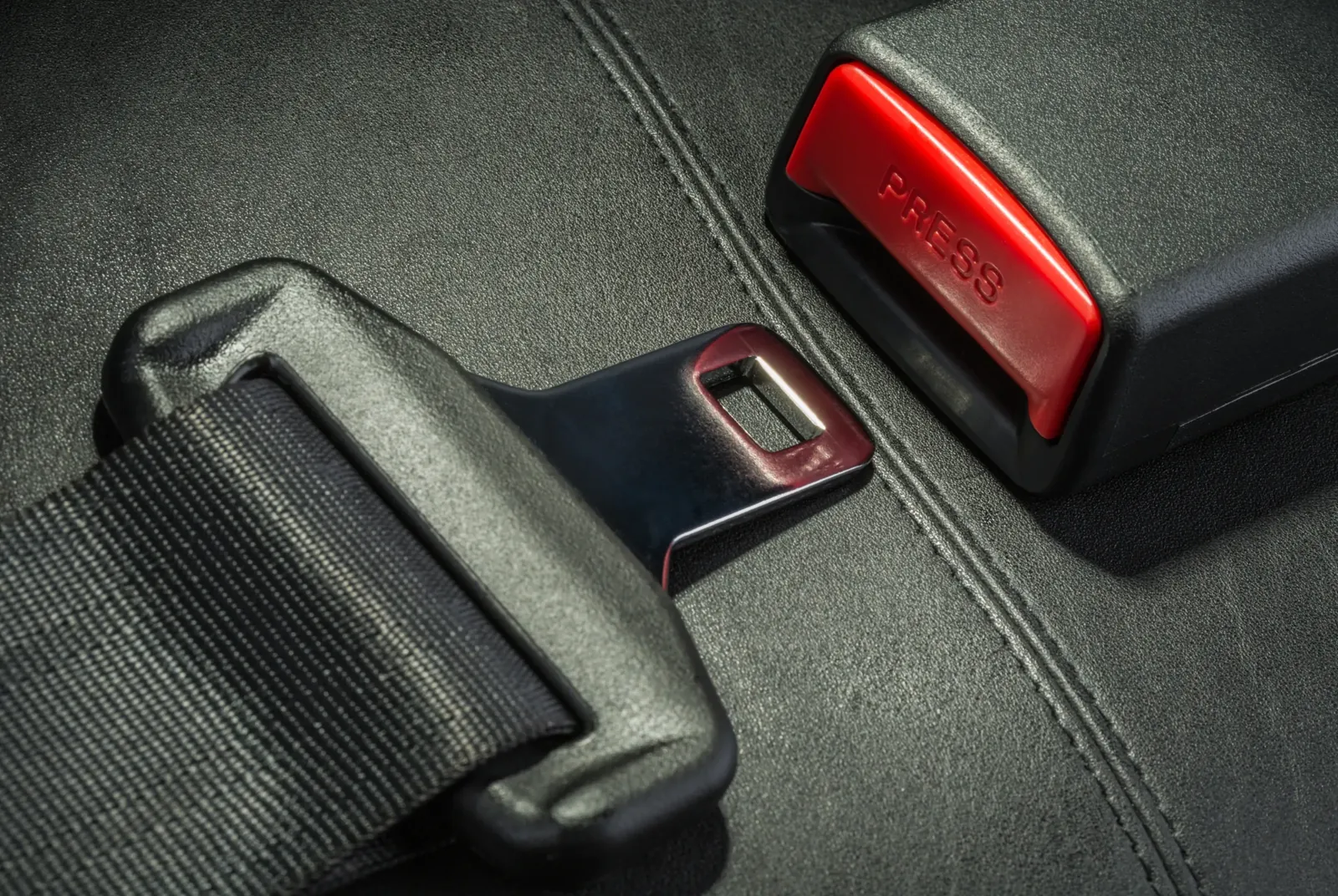 Seat belt buckle body with its red release button, mounted alongside a fabric vehicle seat.