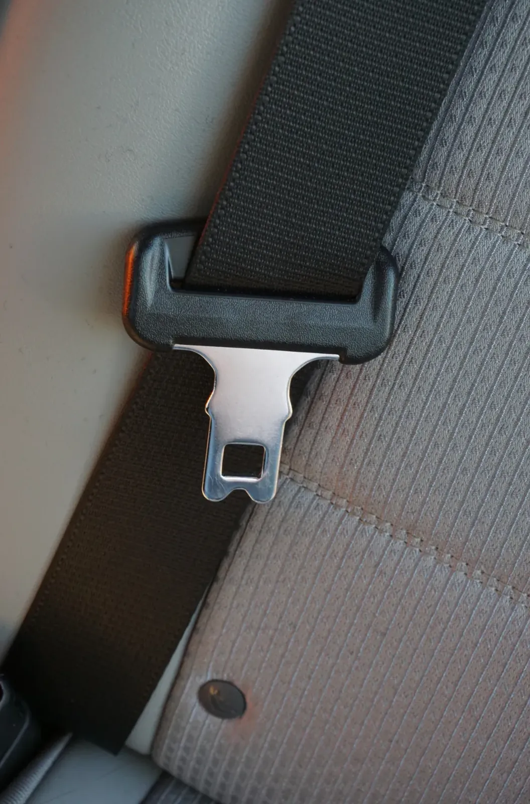 Seat belt tongue plate latched into the buckle beside a light-coloured vehicle seat pillar.